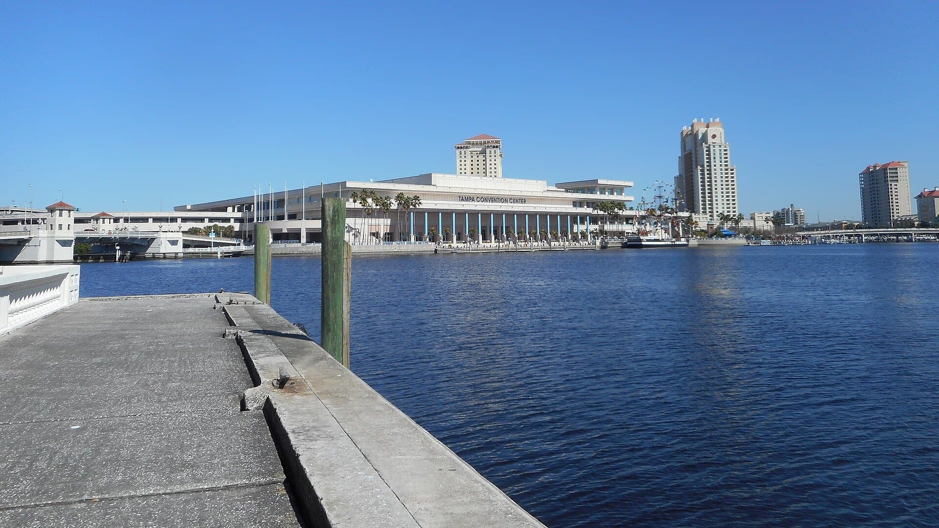 The Tampa Convention Center on the Hillsborough River under a clear blue sky, seen from a seawall along Bayshore Boulevard